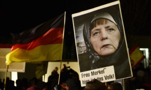A protestor holds a poster with an image of German Chancellor Angela Merkel wearing a headscarf in front of the Reichstag building with a crescent on top during a rally of the group Patriotic Europeans against the Islamization of the West, or PEGIDA, in Dresden, Germany, Monday, Jan. 12, 2015. (AP Photo/Jens Meyer)
